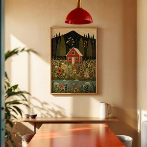 May include: A framed art print depicts a red house nestled in a colorful field of flowers, with a mountain backdrop and a flowing river. The artwork is in a gold frame, hanging on a beige wall. A red pendant light hangs above a wooden table.