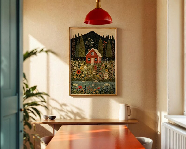 May include: A framed art print depicts a red house nestled in a colorful field of flowers, with a mountain backdrop and a flowing river. The artwork is in a gold frame, hanging on a beige wall. A red pendant light hangs above a wooden table.