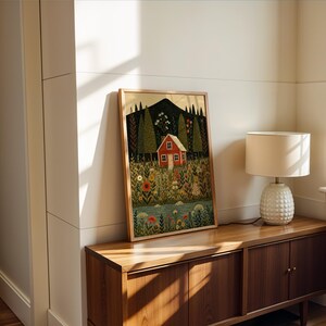 May include: A framed art print featuring a red house nestled in a colorful meadow with a mountain backdrop. The artwork is in a wooden frame and leans against a white wall. A lamp with a white shade sits on a wooden cabinet.