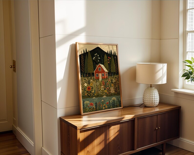 May include: A framed art print featuring a red house nestled in a colorful meadow with a mountain backdrop. The artwork is in a wooden frame and leans against a white wall. A lamp with a white shade sits on a wooden cabinet.