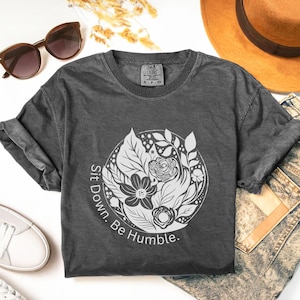 May include: Dark gray t-shirt with a floral design in a circle and the words "Sit Down. Be Humble." printed in white. The shirt is styled with sunglasses, a hat, and jeans.