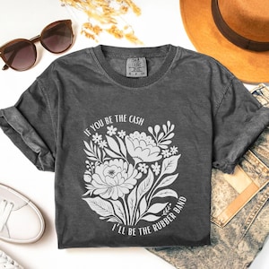 May include: Dark gray t-shirt with a floral design and the text "If you be the cash, I'll be the rubber band." The shirt is styled with sunglasses, a hat, and jeans.