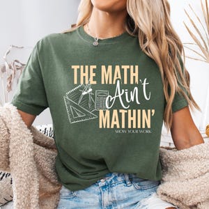 May include: Green t-shirt with the text "THE MATH Ain't MATHIN' SHOW YOUR WORK" in a beige font. The shirt features a graphic of a protractor, calculator, and other math tools.