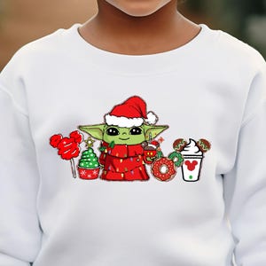 May include: A white sweatshirt featuring a graphic of a green alien character wearing a Santa hat and wrapped in Christmas lights. The graphic includes holiday-themed elements like a candy cane, a Christmas tree, and a coffee cup. The child is smiling.