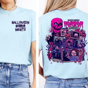 May include: Light blue t-shirt with "HALLOWEEN HORROR NIGHTS" text on the front and a graphic on the back featuring a pink skull, horror movie characters, and vintage televisions. The shirt is paired with blue jeans.