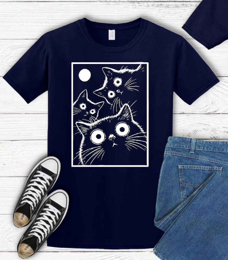 May include: Navy blue t-shirt featuring a white graphic of four cartoon cats with large eyes and a moon. The cats are inside a white rectangle. Black and white sneakers and blue jeans are also in the image.