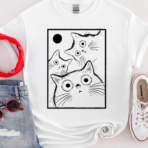 May include: White t-shirt featuring a black and white graphic of three cats with large, surprised eyes. The cats are inside a black rectangle with a black circle in the upper left corner. The shirt is paired with denim shorts and white sneakers.