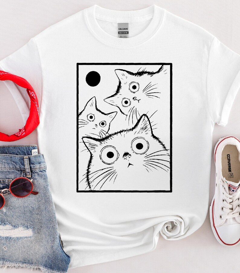 May include: White t-shirt featuring a black and white graphic of three cats with large, surprised eyes. The cats are inside a black rectangle with a black circle in the upper left corner. The shirt is paired with denim shorts and white sneakers.