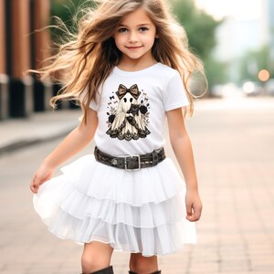 Pu&ograve; includere: Una t-shirt bianca a maniche corte con la grafica di un fantasma che indossa un fiocco leopardato e tiene in braccio un gatto nero, circondato da rose e piccoli fiocchi. Abbinata a una gonna in tulle bianco a balze e una cintura scura.
