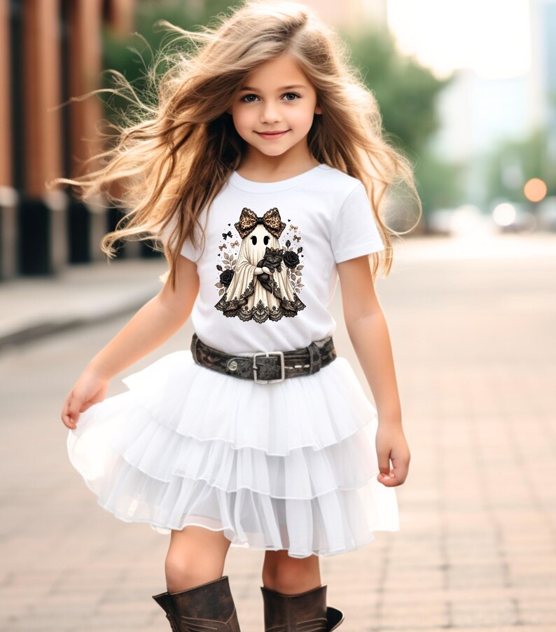 Pu&ograve; includere: Una t-shirt bianca a maniche corte con la grafica di un fantasma che indossa un fiocco leopardato e tiene in braccio un gatto nero, circondato da rose e piccoli fiocchi. Abbinata a una gonna in tulle bianco a balze e una cintura scura.