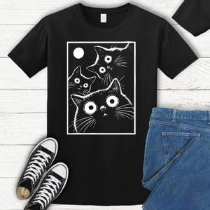 May include: Black t-shirt featuring a white graphic of four cartoon cats with large eyes and a moon. The cats are inside a white rectangle. A pair of black and white sneakers and blue jeans are also visible.