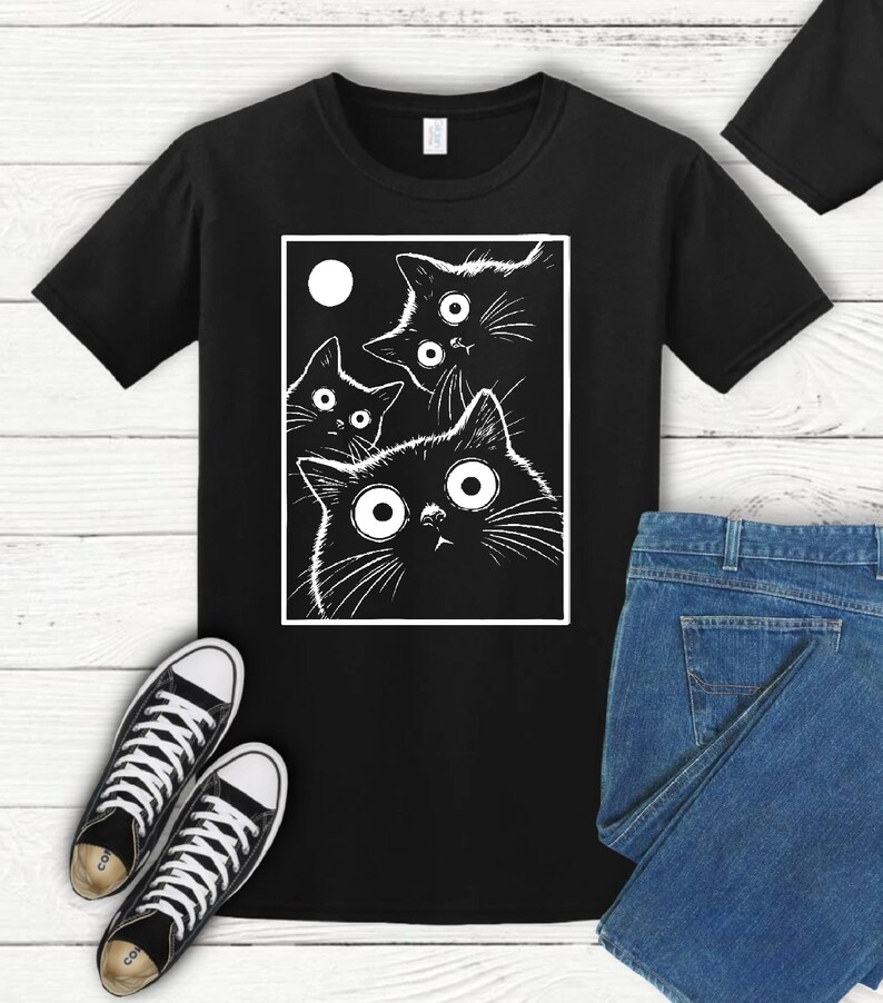 May include: Black t-shirt featuring a white graphic of four cartoon cats with large eyes and a moon. The cats are inside a white rectangle. A pair of black and white sneakers and blue jeans are also visible.