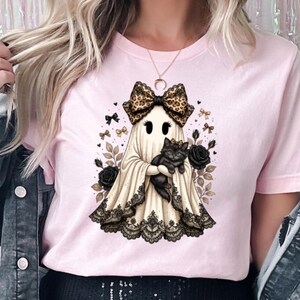 Pu&ograve; includere: Una t-shirt rosa chiaro con la grafica di un fantasma che indossa un fiocco leopardato e tiene in braccio un gatto nero. Il fantasma &egrave; circondato da rose nere, piccoli fiocchi e foglie, con un bordo in pizzo nero sul fondo del lenzuolo.