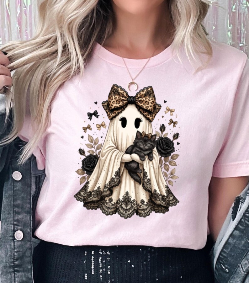 Pu&ograve; includere: Una t-shirt rosa chiaro con la grafica di un fantasma che indossa un fiocco leopardato e tiene in braccio un gatto nero. Il fantasma &egrave; circondato da rose nere, piccoli fiocchi e foglie, con un bordo in pizzo nero sul fondo del lenzuolo.
