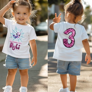May include: A white t-shirt with a colorful graphic of a spider web and a superhero character. The text "Birthday Girl Ryli" is below the graphic. The back of the shirt has a large, sparkly pink number "3". The shirt is paired with denim shorts.