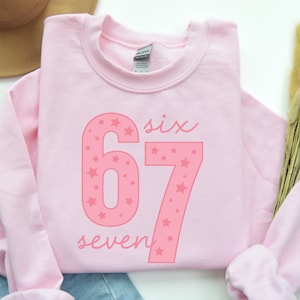 May include: A light pink sweatshirt with the numbers "67" in a darker pink, filled with star shapes. The word "six" is above the number 6 and the word "seven" is below the number 7. The sweatshirt is folded.