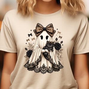 Pu&ograve; includere: Una t-shirt beige con la grafica di un fantasma bianco che indossa un grande fiocco leopardato e tiene in braccio un gattino nero. Il fantasma &egrave; decorato con un bordo in pizzo nero sull'orlo ed &egrave; circondato da rose nere, fiocchetti e foglie.