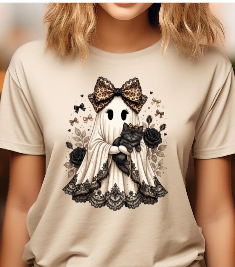 Pu&ograve; includere: Una t-shirt beige con la grafica di un fantasma bianco che indossa un grande fiocco leopardato e tiene in braccio un gattino nero. Il fantasma &egrave; decorato con un bordo in pizzo nero sull'orlo ed &egrave; circondato da rose nere, fiocchetti e foglie.