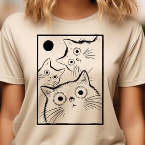 May include: Beige t-shirt featuring a black and white illustration of three cats with large, surprised eyes and a black circle. The cats are inside a black rectangular frame. The shirt has short sleeves.