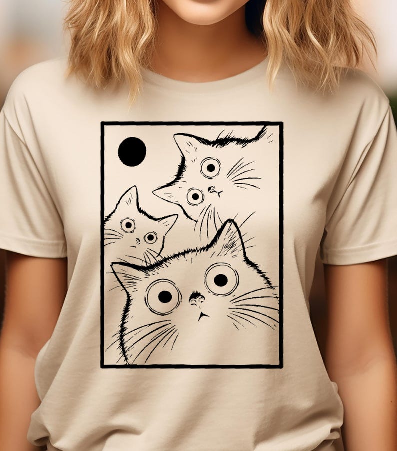 May include: Beige t-shirt featuring a black and white illustration of three cats with large, surprised eyes and a black circle. The cats are inside a black rectangular frame. The shirt has short sleeves.
