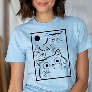 May include: Light blue t-shirt featuring a black and white graphic of three cartoon cats with large eyes and a black circle. The cats are inside a black rectangle. The shirt has short sleeves.