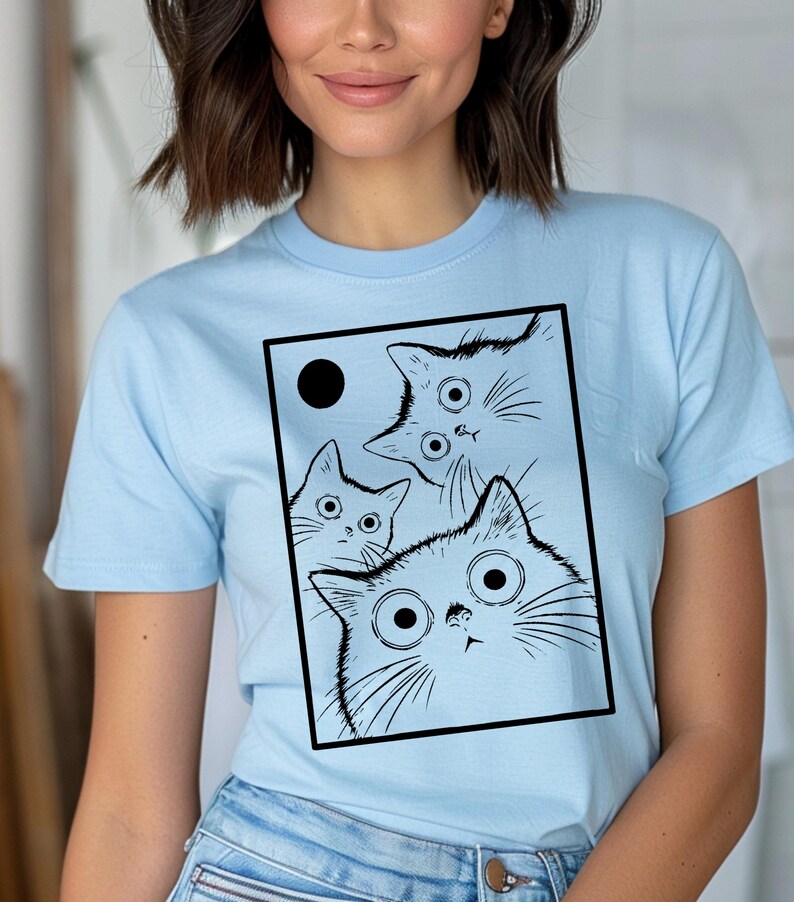 May include: Light blue t-shirt featuring a black and white graphic of three cartoon cats with large eyes and a black circle. The cats are inside a black rectangle. The shirt has short sleeves.