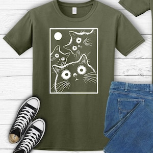 May include: Olive green t-shirt featuring a white graphic of four cats with large, surprised eyes and a moon. The cats are inside a white rectangular frame. Black and white sneakers and blue jeans are also visible.