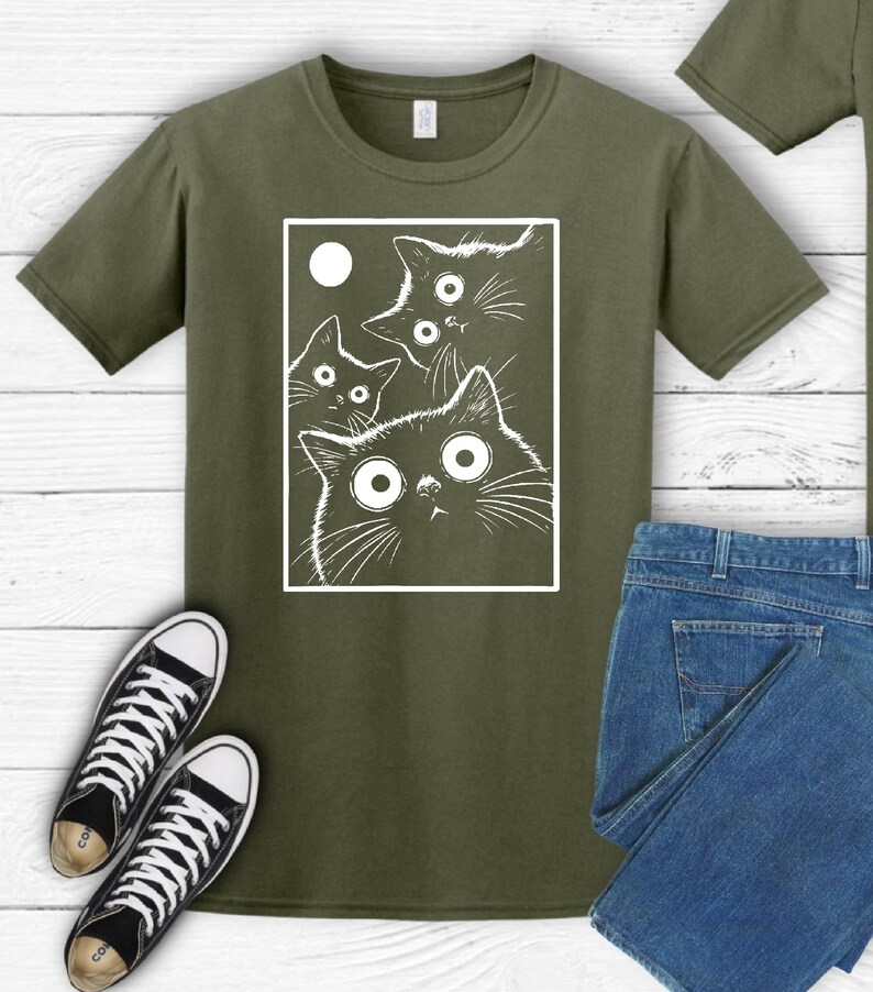 May include: Olive green t-shirt featuring a white graphic of four cats with large, surprised eyes and a moon. The cats are inside a white rectangular frame. Black and white sneakers and blue jeans are also visible.