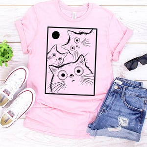 May include: A light pink t-shirt featuring a black and white illustration of four cats with large eyes. The cats are inside a black rectangle with a black circle in the upper left corner. The shirt is paired with denim shorts and white sneakers.