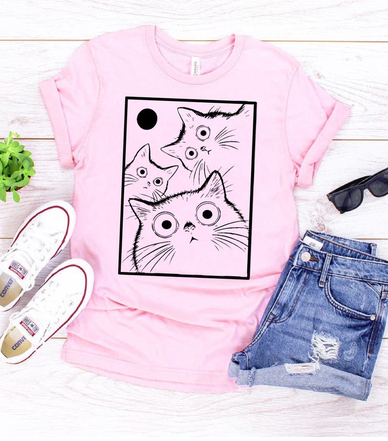 May include: A light pink t-shirt featuring a black and white illustration of four cats with large eyes. The cats are inside a black rectangle with a black circle in the upper left corner. The shirt is paired with denim shorts and white sneakers.