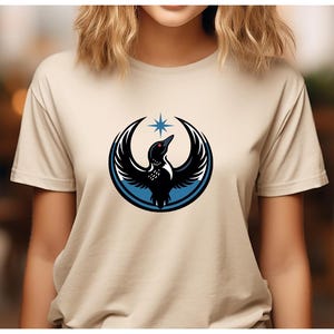 May include: Beige t-shirt featuring a black and white bird design with a blue outline and a star above it. The bird has its wings spread, and the design is centered on the shirt. The shirt is made of a soft material.