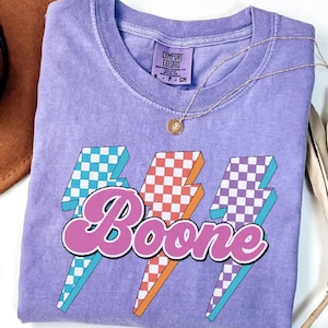 May include: A lavender Comfort Colors t-shirt with a graphic design featuring three lightning bolts with checkered patterns in blue, orange, and purple. The word "Boone" is written in a script font across the lightning bolts. A gold necklace is draped across the shirt.