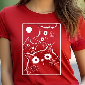 May include: Red t-shirt featuring a white graphic of four cartoon cats with wide eyes and a white circle. The cats are inside a white rectangular frame. The t-shirt has short sleeves.