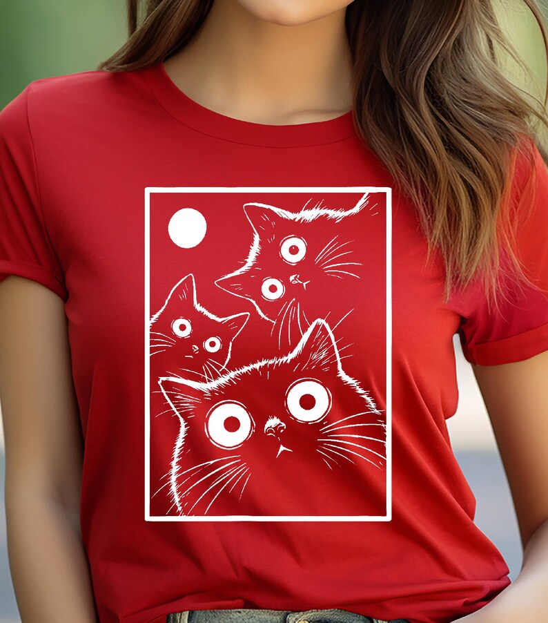 May include: Red t-shirt featuring a white graphic of four cartoon cats with wide eyes and a white circle. The cats are inside a white rectangular frame. The t-shirt has short sleeves.