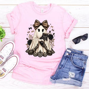 Pu&ograve; includere: Una t-shirt rosa chiaro con la grafica di un fantasma che indossa un fiocco leopardato e tiene in braccio un gatto nero. Il fantasma &egrave; decorato con un bordo in pizzo nero sull'orlo ed &egrave; circondato da rose nere, fiocchetti e cuori.