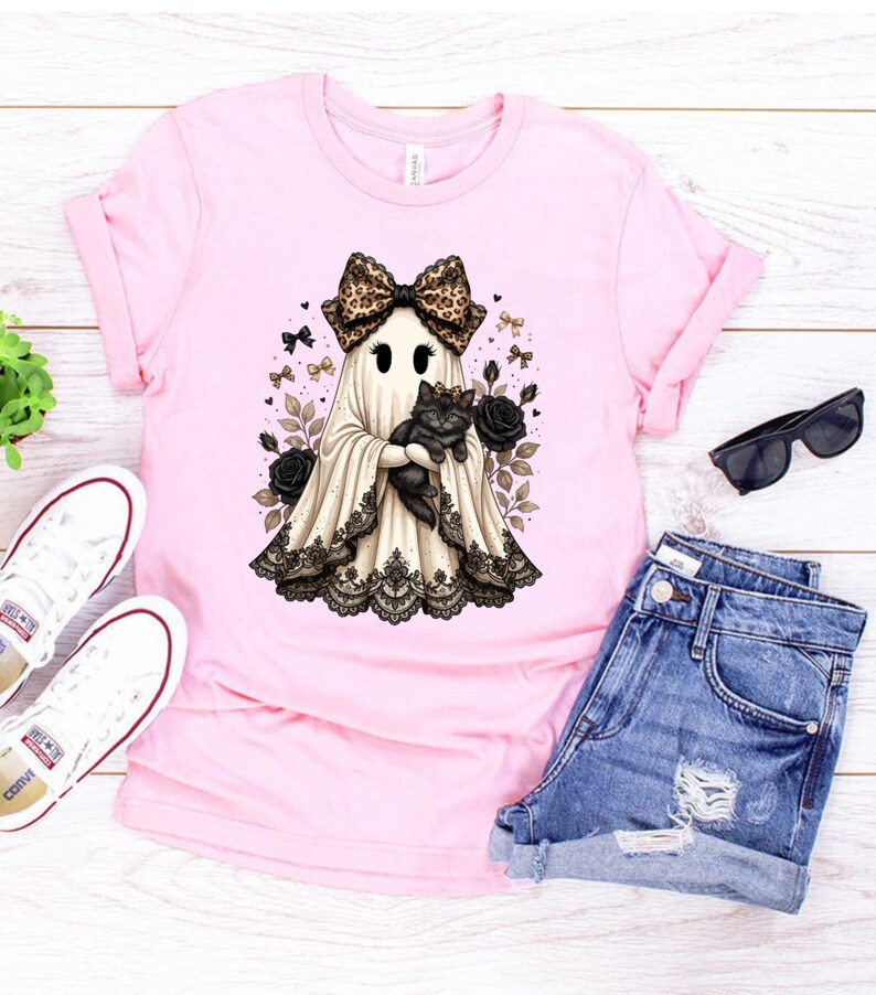 Pu&ograve; includere: Una t-shirt rosa chiaro con la grafica di un fantasma che indossa un fiocco leopardato e tiene in braccio un gatto nero. Il fantasma &egrave; decorato con un bordo in pizzo nero sull'orlo ed &egrave; circondato da rose nere, fiocchetti e cuori.