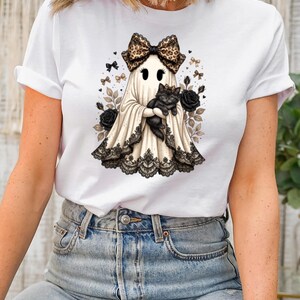 Pu&ograve; includere: Una t-shirt bianca a maniche corte con la grafica di un fantasma che indossa un fiocco leopardato e tiene in braccio un gatto nero. Il fantasma &egrave; decorato con un bordo in pizzo nero sull'orlo, circondato da rose nere e piccoli fiocchi.