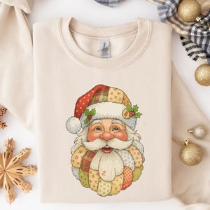 May include: Cream-colored sweatshirt featuring a Santa Claus graphic. The Santa illustration has a patchwork design on his hat and beard, with red, green, and yellow accents. The Santa Claus graphic is centered on the front of the sweatshirt.