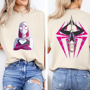 Puede incluir: Camiseta beige con un dise&ntilde;o de superhero&iacute;na en la parte delantera, con una m&aacute;scara con capucha blanca y rosa y un traje negro y blanco. La parte trasera de la camiseta muestra a la misma superhero&iacute;na colgada boca abajo en forma de diamante rosa y azul.