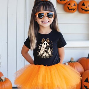 Pu&ograve; includere: Una t-shirt nera a maniche corte con la grafica di un fantasma che indossa un fiocco leopardato e tiene in braccio un gatto nero, abbinata a una gonna tutu in tulle arancione brillante. Il design del fantasma presenta dettagli in pizzo nero.