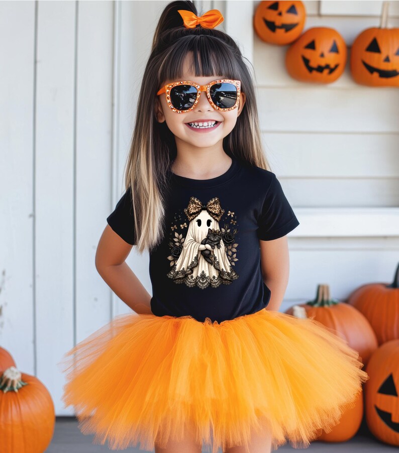 Pu&ograve; includere: Una t-shirt nera a maniche corte con la grafica di un fantasma che indossa un fiocco leopardato e tiene in braccio un gatto nero, abbinata a una gonna tutu in tulle arancione brillante. Il design del fantasma presenta dettagli in pizzo nero.