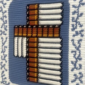 May include: Crocheted wall hanging featuring a cross design. The cross is brown and white, with a blue background. The border is white with blue floral accents. The piece is handmade and has a textured appearance.