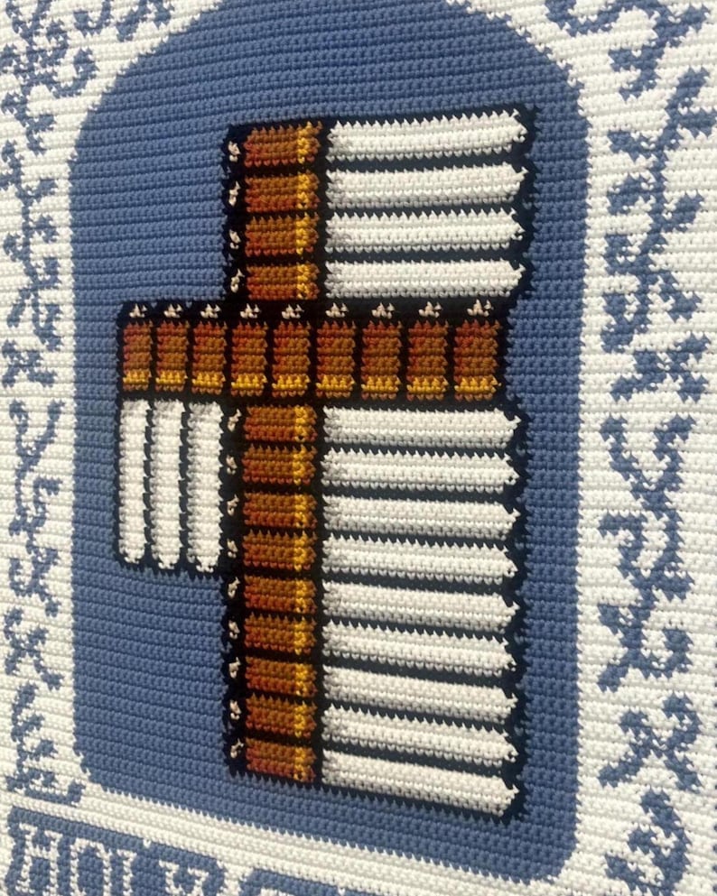 May include: Crocheted wall hanging featuring a cross design. The cross is brown and white, with a blue background. The border is white with blue floral accents. The piece is handmade and has a textured appearance.