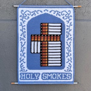 Könnte beinhalten: Blau-weiße gehäkelte Wanddekoration mit einem Kreuz aus Zigaretten. Das Kreuz befindet sich in der Mitte, umgeben von einem blauen Bogen. Der Schriftzug "HOLY SMOKES" befindet sich unten.