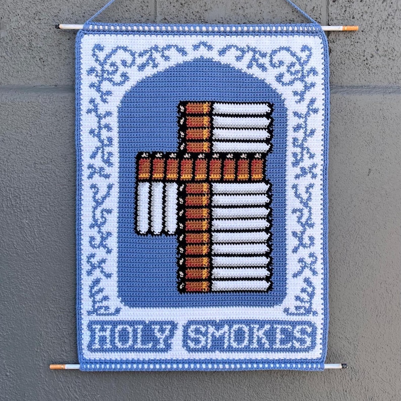 May include: A blue and white crocheted wall hanging with a cross made of cigarettes. The cross is in the center, with a blue arch shape around it. The words "HOLY SMOKES" are at the bottom.