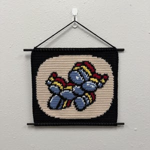 May include: A handmade, crocheted wall hanging featuring a pixelated design of a blue, red, and yellow creature. The artwork is set against a beige background within a black border, suspended by a black rod and string.