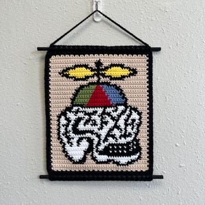 May include: Crocheted wall hanging with a colourful design. The artwork depicts a stylized skull with a propeller, using black, white, red, blue, green, and yellow yarn. It's framed with a black border and hanging rod.
