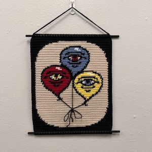 May include: Crocheted wall hanging featuring three balloon shapes in red, blue, and yellow, each with an eye design. The balloons are set against a beige background with a black border and hanging rod.