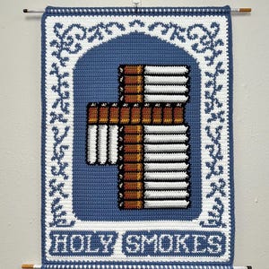 May include: A handmade, crocheted wall hanging with a blue background and a white border. The design features a cross made of cigarettes, with the text "HOLY SMOKES" at the bottom. The piece is hung with two dowels.