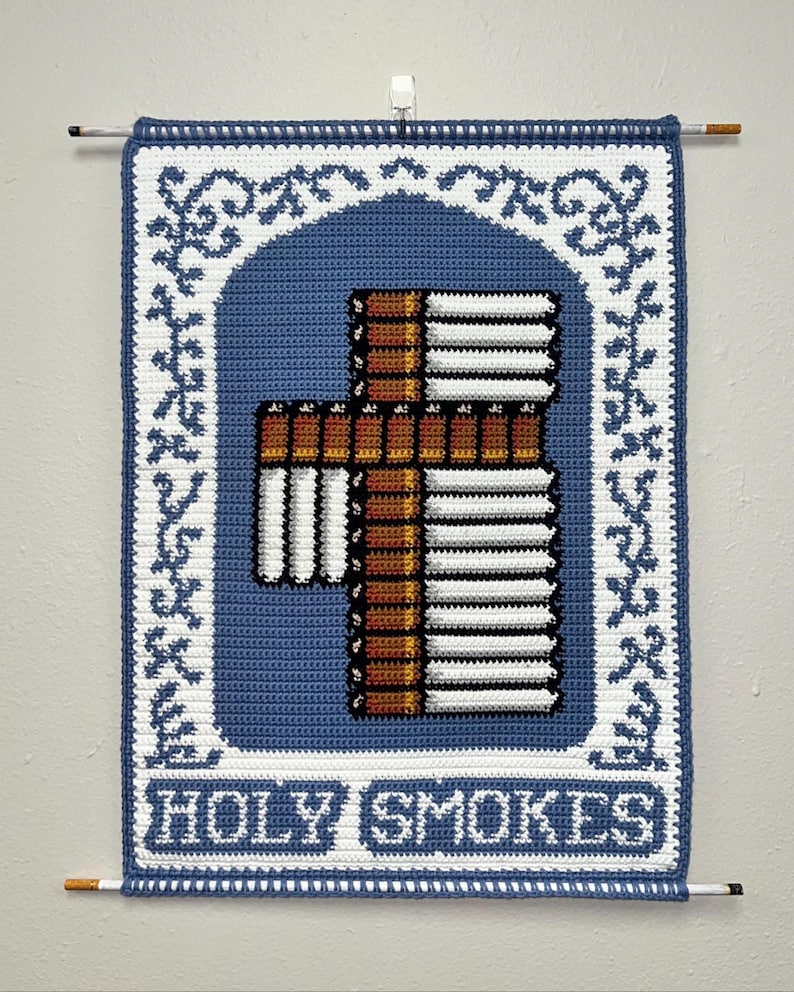 May include: A handmade, crocheted wall hanging with a blue background and a white border. The design features a cross made of cigarettes, with the text "HOLY SMOKES" at the bottom. The piece is hung with two dowels.